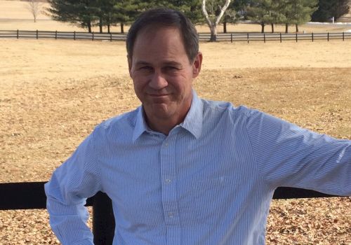 A man stands by a fence in a field on a sunny day, with trees and a clear sky in the background. He wears a light shirt and smiles at the camera.