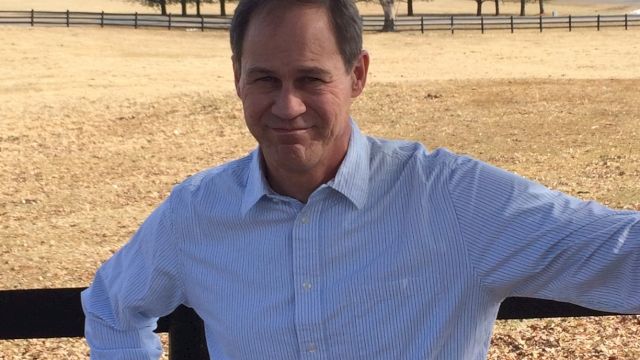 A man stands by a fence in a field on a sunny day, with trees and a clear sky in the background. He wears a light shirt and smiles at the camera.