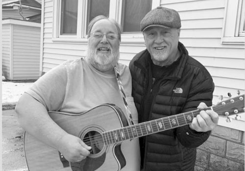 Two men smiling, with one holding a guitar and the other wearing a cap, standing in front of a house.