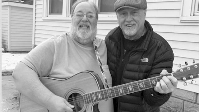 Two men smiling, with one holding a guitar and the other wearing a cap, standing in front of a house.