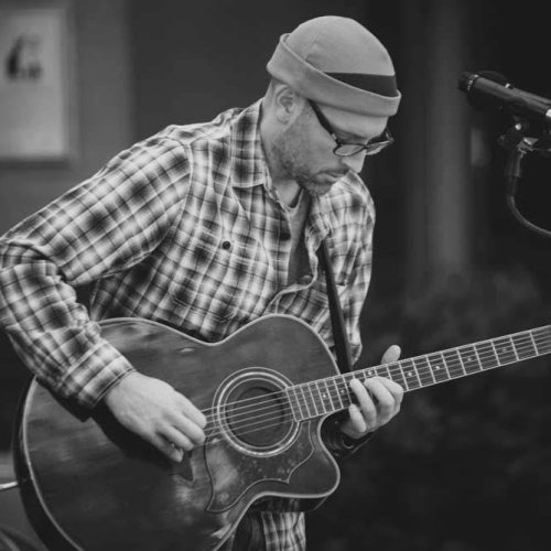 A person in a plaid shirt and beanie playing an acoustic guitar in front of a microphone.