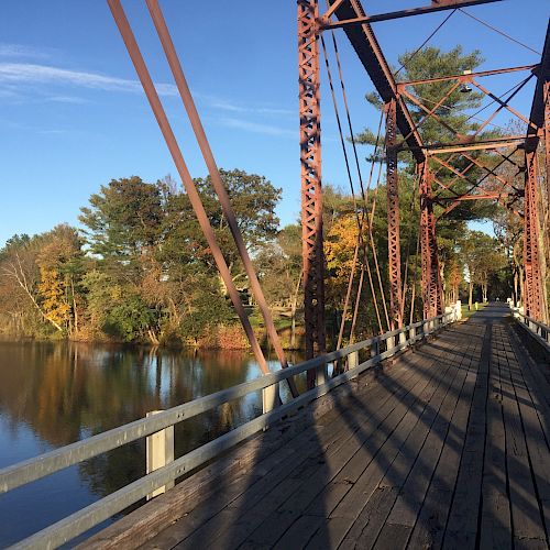 A picturesque wooden bridge with rusted metal railings extends over a calm river surrounded by trees, bathed in sunlight under a clear blue sky.