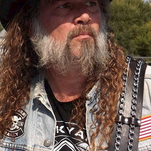 A bearded man with long hair is wearing a black hat and denim vest adorned with patches and a leather strap with chains ending the sentence.