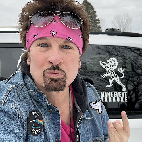 A person in a pink bandana with sunglasses poses in front of a vehicle with a logo that says "MAIN EVENT KARAOKE."