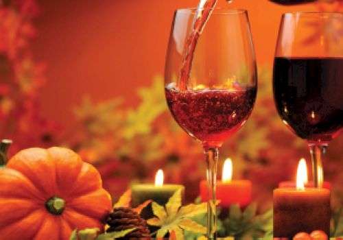 The image shows two glasses of wine, pumpkins, candles, pinecones, and autumn leaves, creating a warm, festive, and cozy autumn atmosphere.
