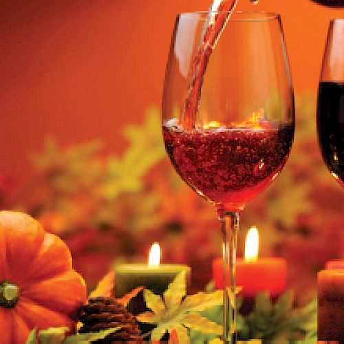 The image shows two glasses of wine, pumpkins, candles, pinecones, and autumn leaves, creating a warm, festive, and cozy autumn atmosphere.