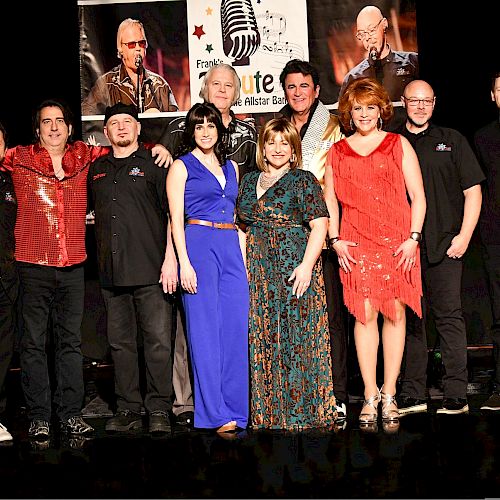 A group of people standing on a stage, with a backdrop featuring microphones and performers, wearing a variety of outfits.