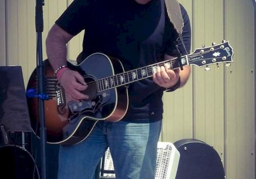 A person is playing an acoustic guitar on a stage with a microphone stand nearby. They are wearing a black shirt and cap, and blue jeans.