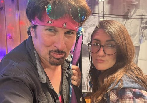 Two people posing for a photo with colorful lighting in the background. One person wears a headband, and the other has glasses and a plaid shirt.