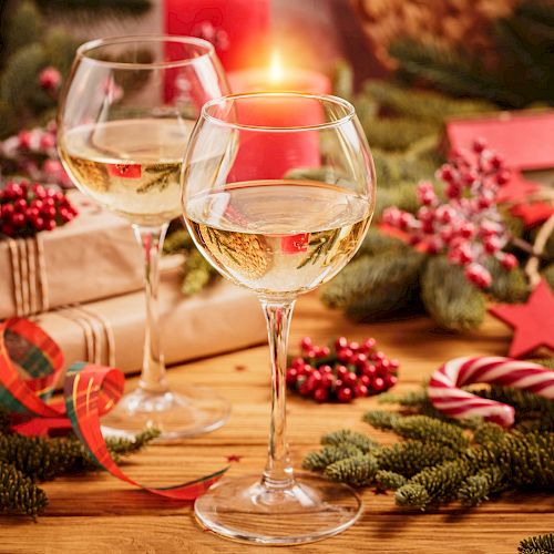 Two wine glasses, wrapped gifts, and festive decorations, including pine branches, berries, a candy cane, and ribbon.