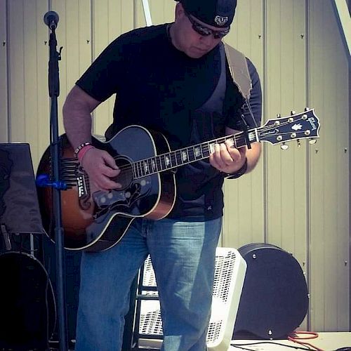 A person wearing a black shirt and cap plays an acoustic guitar on an outdoor stage with a mic stand and music equipment nearby.