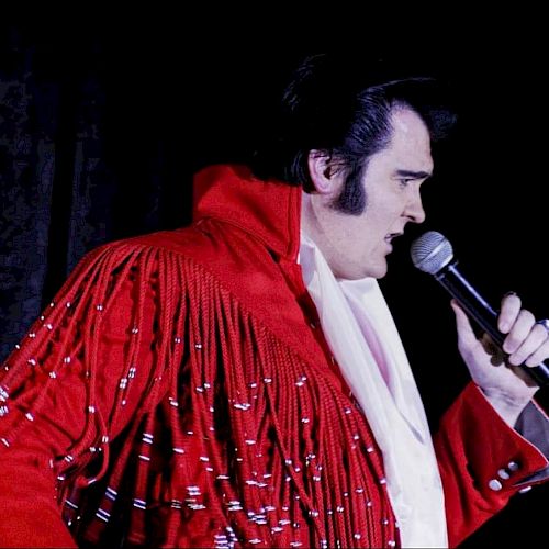 A person wearing a red, fringed jacket is holding a microphone and appears to be singing or performing against a dark background.