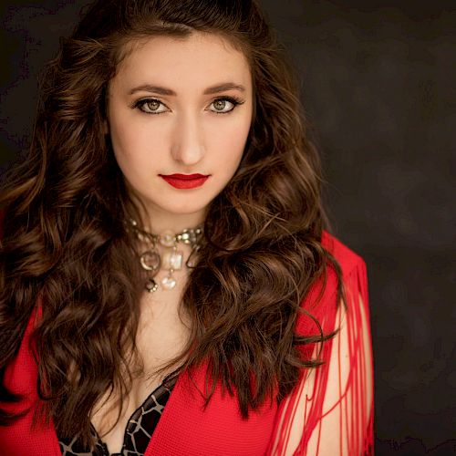 A woman with long, wavy hair wearing a red outfit and a necklace, against a dark background.