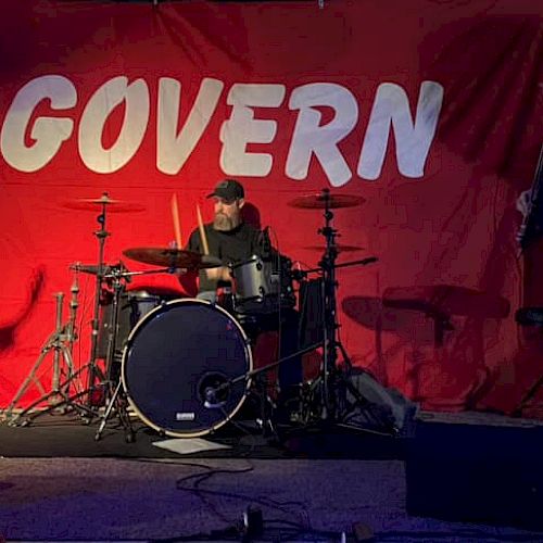 Three musicians performing on stage with instruments; a red banner behind them reads 