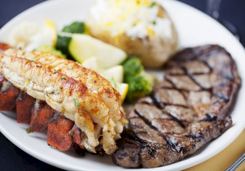 The image shows a plate of surf and turf with a grilled lobster tail, a steak, baked potato, and steamed vegetables including broccoli and lemon wedges.