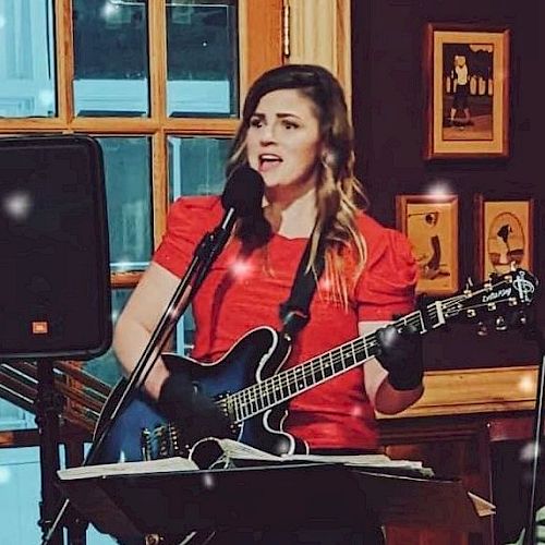 A woman in a red dress is playing an electric guitar and singing into a microphone at an indoor venue with framed pictures on the wall behind.