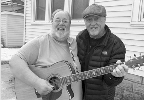 Two smiling men outside a house, one holding a guitar, the other with an arm around his shoulder, both appearing happy and friendly.