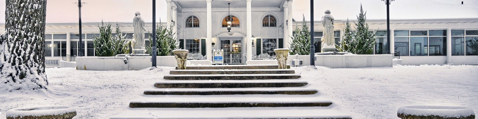 A snow-covered staircase leads to a grand, white two-story building with a balcony and street lamps lining the path.