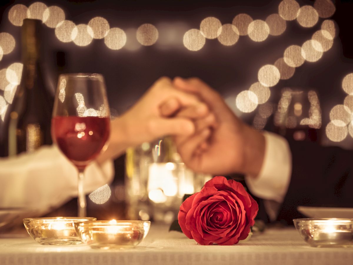 A romantic setting with two people holding hands across a candlelit table, featuring a red rose and wine glasses.