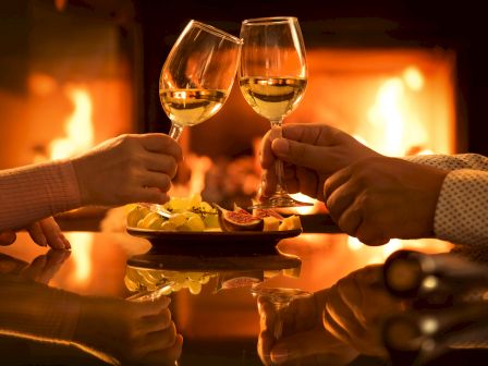 Two people clink glasses of white wine near a cozy fireplace, with a plate of lemon slices and fruit on the table.
