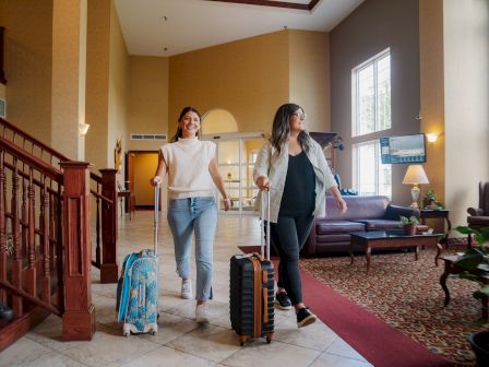 Two women are walking through a hotel lobby with rolling suitcases, appearing cheerful as they move towards the exit.