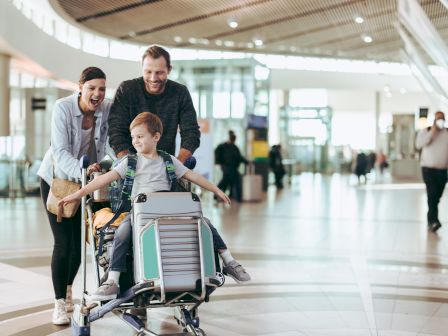 A family is at an airport. A child is seated on a luggage cart, being pushed by an adult, while another adult joyfully walks beside them.