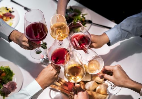 A group of people toasting with glasses of red and white wine at a table set with food, including salads and bread, from an overhead perspective.