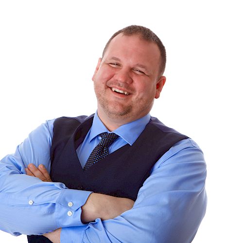 The image shows a smiling man in a blue shirt and dark vest, with his arms crossed, against a plain white background.