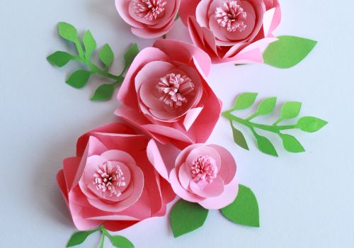 The image shows pink paper flowers with green leaves arranged on a light background. The flowers and leaves appear to be crafted from paper.