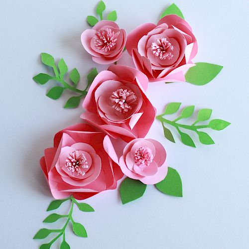 The image shows pink paper flowers with green leaves arranged on a light background. The flowers and leaves appear to be crafted from paper.