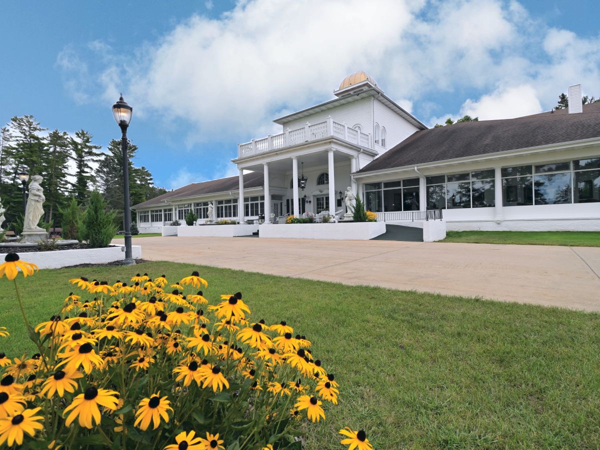 A large white building with columns, surrounded by green grass and yellow flowers, under a blue sky with clouds.