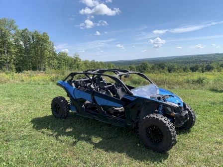 A blue off-road vehicle is parked on a grassy field with a scenic forested backdrop under a partly cloudy sky, ending the sentence.