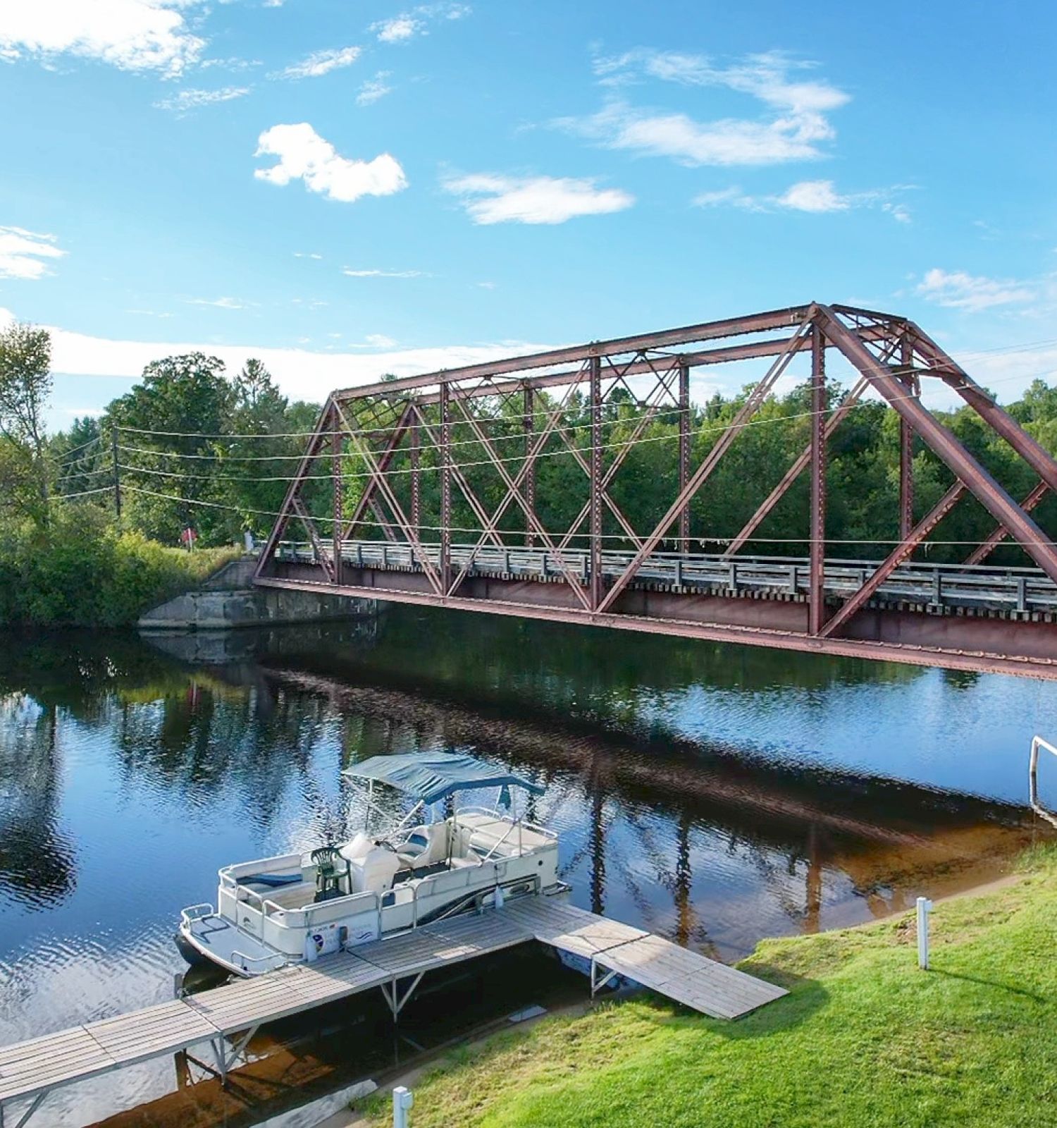 A boat docked near a river with a red truss bridge in the background, surrounded by trees and a clear blue sky ending the sentence.