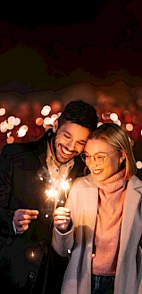 A couple happily holds a sparkler at night, with festive lights blurred in the background.