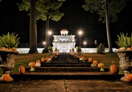 A nighttime scene with a lit building and a staircase lined with pumpkins and plants, leading up to the entrance.