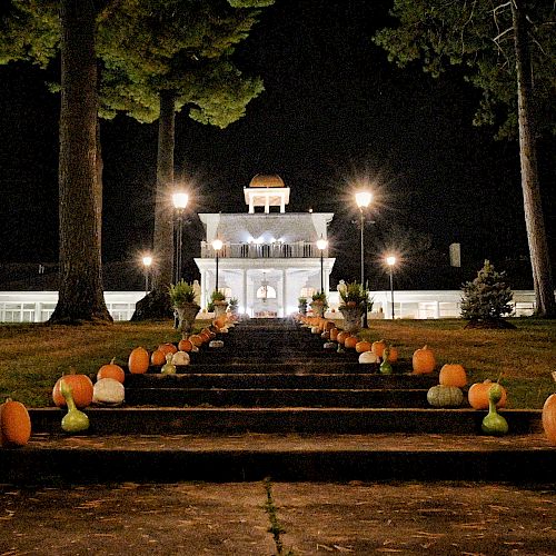 A nighttime scene with a lit building and a staircase lined with pumpkins and plants, leading up to the entrance.