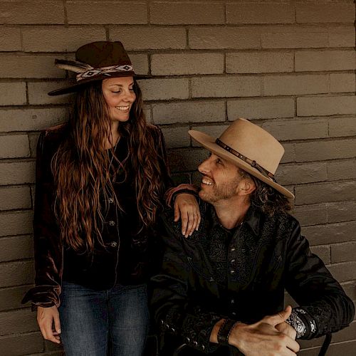 A man and a woman wearing hats are smiling at each other in front of a brick wall.