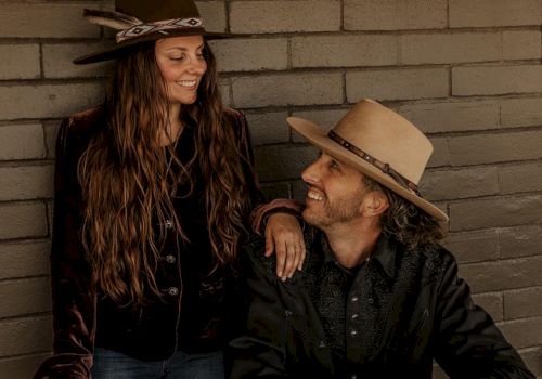 A man and a woman wearing hats are smiling at each other in front of a brick wall.