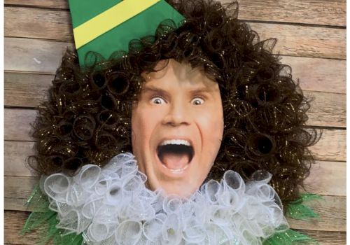 A humorous wreath featuring a person's face with a curly wig, elf hat, and mesh ruffle collar on a wooden background.