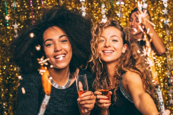 People celebrating with sparklers and drinks in front of a glittery backdrop, looking joyful and festive at a party.