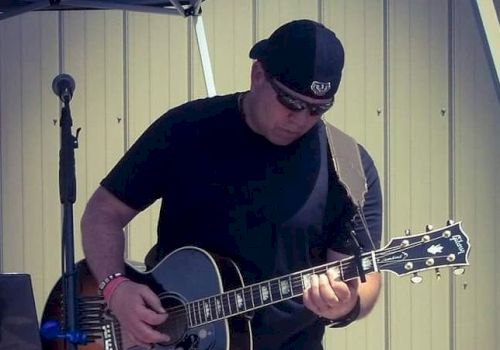 A person wearing sunglasses and a hat is playing an acoustic guitar with a microphone stand beside them in an outdoor setting.