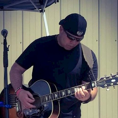 A person wearing sunglasses and a hat is playing an acoustic guitar with a microphone stand beside them in an outdoor setting.