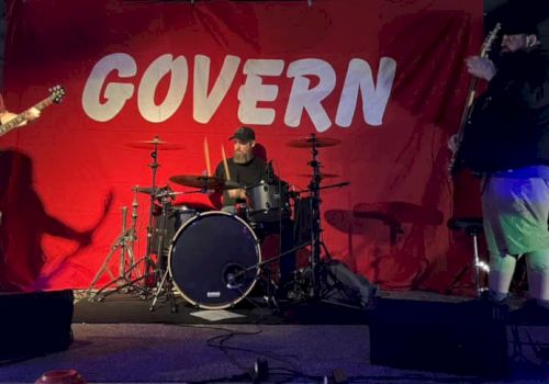 Three people are performing on stage with musical instruments in front of a red backdrop that says "GOVERN".