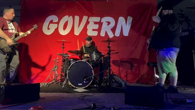 Three people are performing on stage with musical instruments in front of a red backdrop that says "GOVERN".