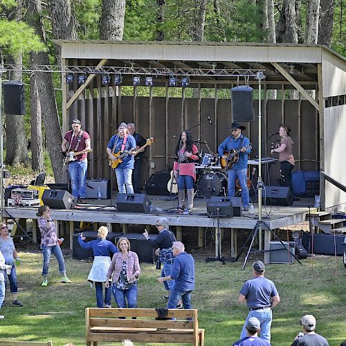 A band performing on an outdoor stage surrounded by a forest, with an audience watching and dancing on the grass.