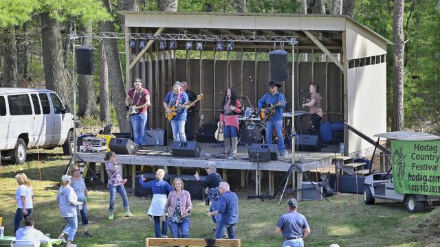 A band performing on an outdoor stage surrounded by a forest, with an audience watching and dancing on the grass.