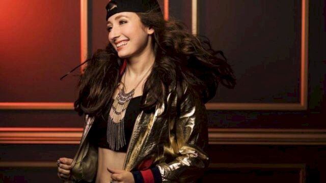 A person smiling, wearing a black cap backwards, a gold and black jacket, layered necklaces, and a crop top, standing against a wall with geometric accents.