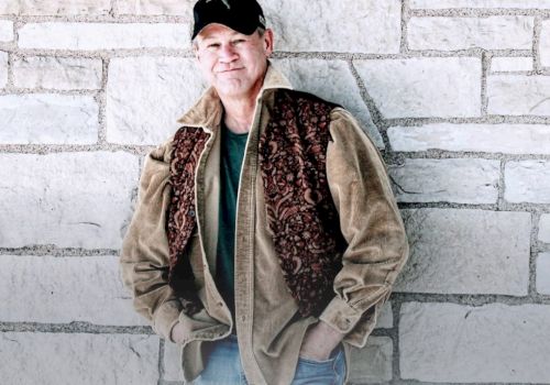 A person wearing a cap, brown jacket, and jeans leans against a brick wall, with hands in pockets, looking relaxed.