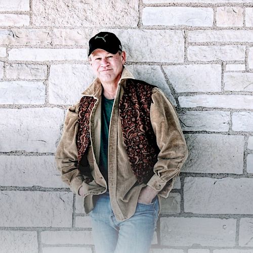 A person wearing a cap, brown jacket, and jeans leans against a brick wall, with hands in pockets, looking relaxed.