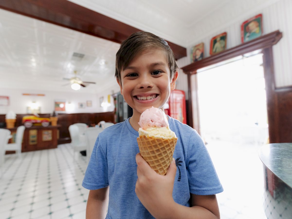 A smiling child in a blue shirt holds an ice cream cone inside a bright, vintage-style ice cream parlor with tiled floors.